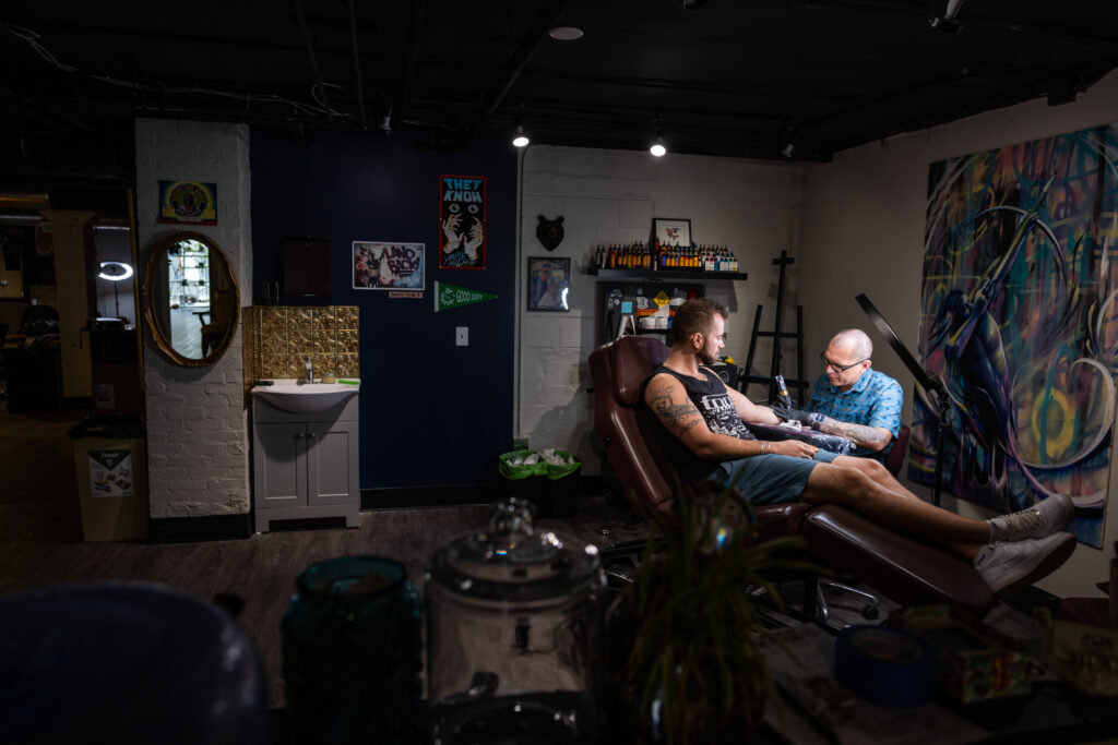 A person tattooing another person sitting on a chair beside them.