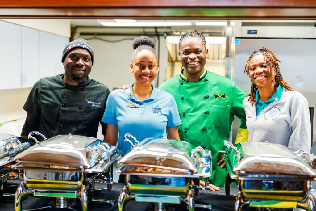 Four smiling people standing in a kitchen behind several covered platters of food.