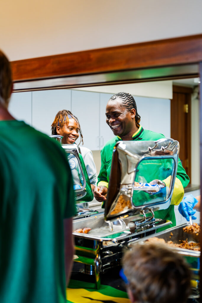 Two smiling people serving food from metal trays in a kitchen.