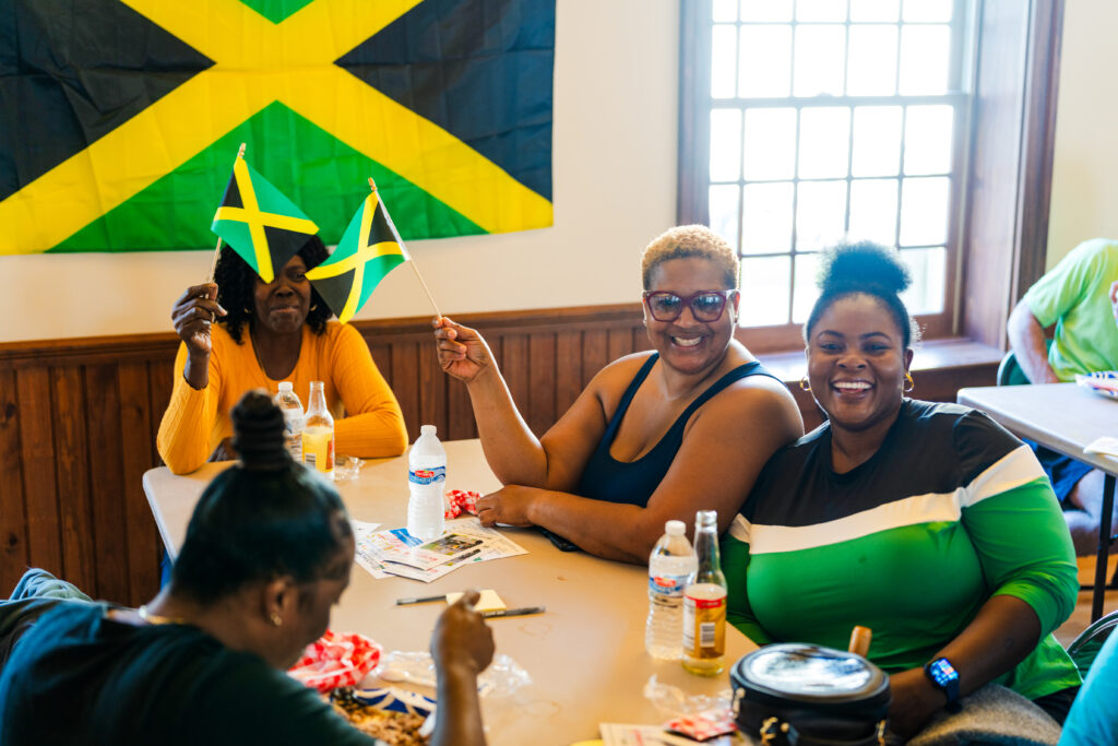 Three smiling people sitting around a table holding small Jamaican flags, with a larger Jamaican flag hanging behind them.