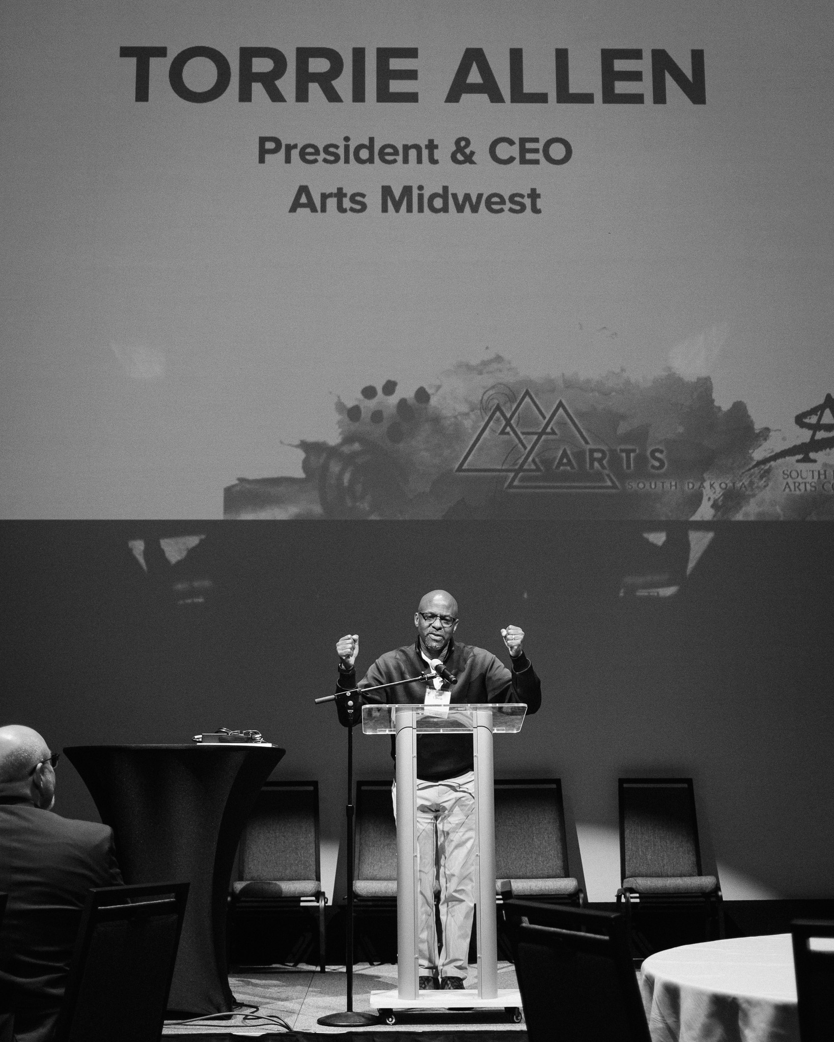 A black and white photo of a Black man at a podium raising both first. Text in the background reads Torrie Allen President + CEO Arts Midwest