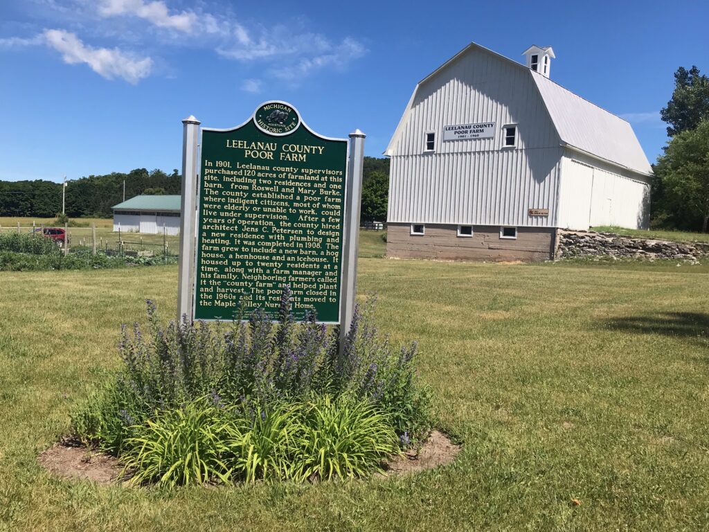 An all-white barn at a distance with a historic marked in the front of it. The marker is titled "Leelanau County Poor Farm"