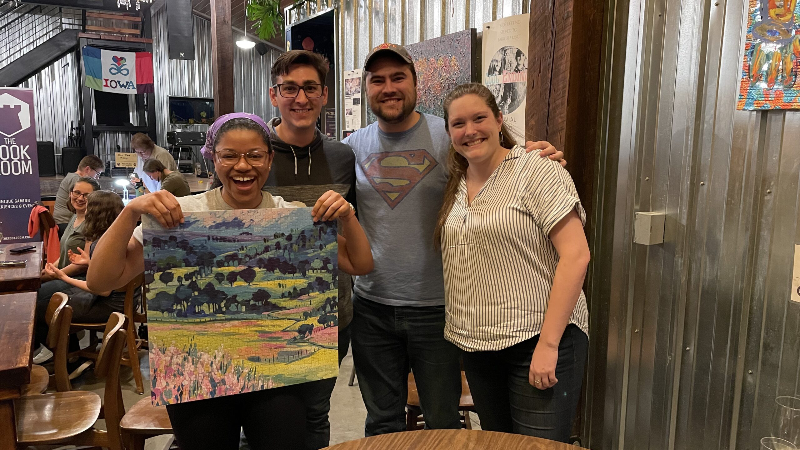 A group of four people stand together and smile. One of them is holding up a completed jigsaw puzzle.