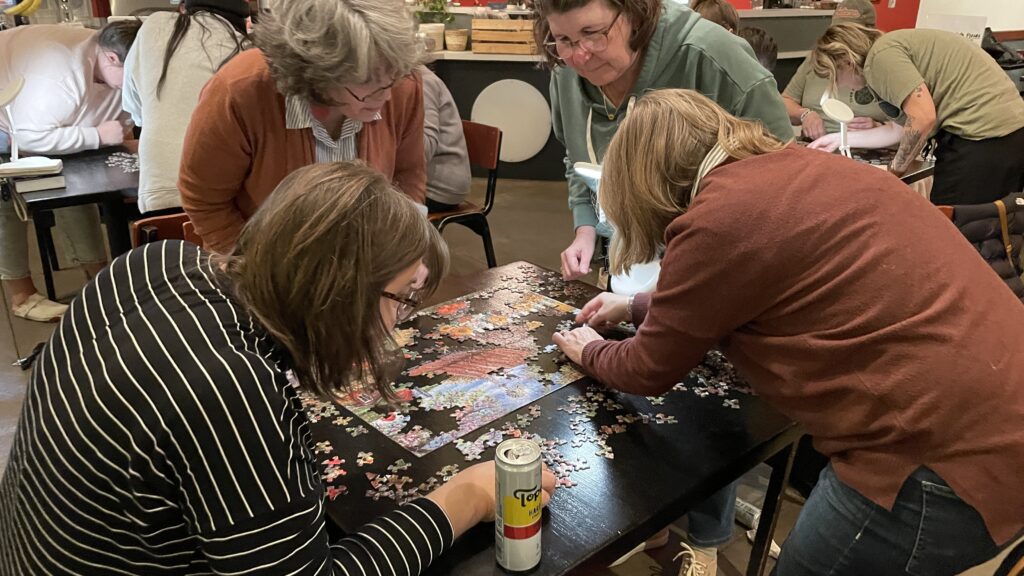 A groups of people hunched over a table as they solve a jigsaw puzzle.