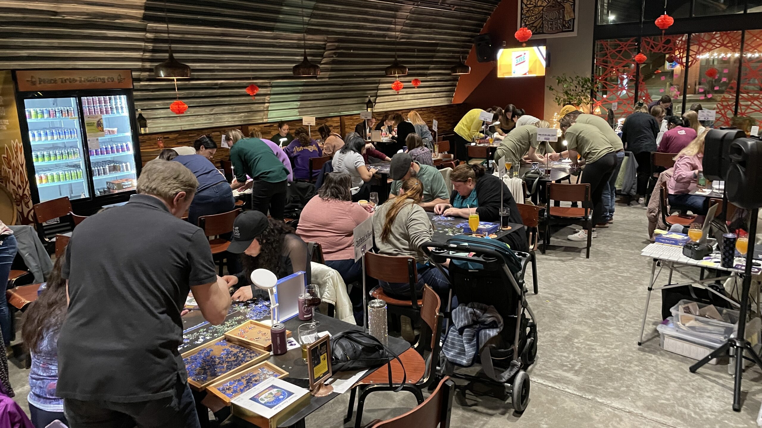 Groups of people stand around tables in a large room as they work on solving their jigsaw puzzles.