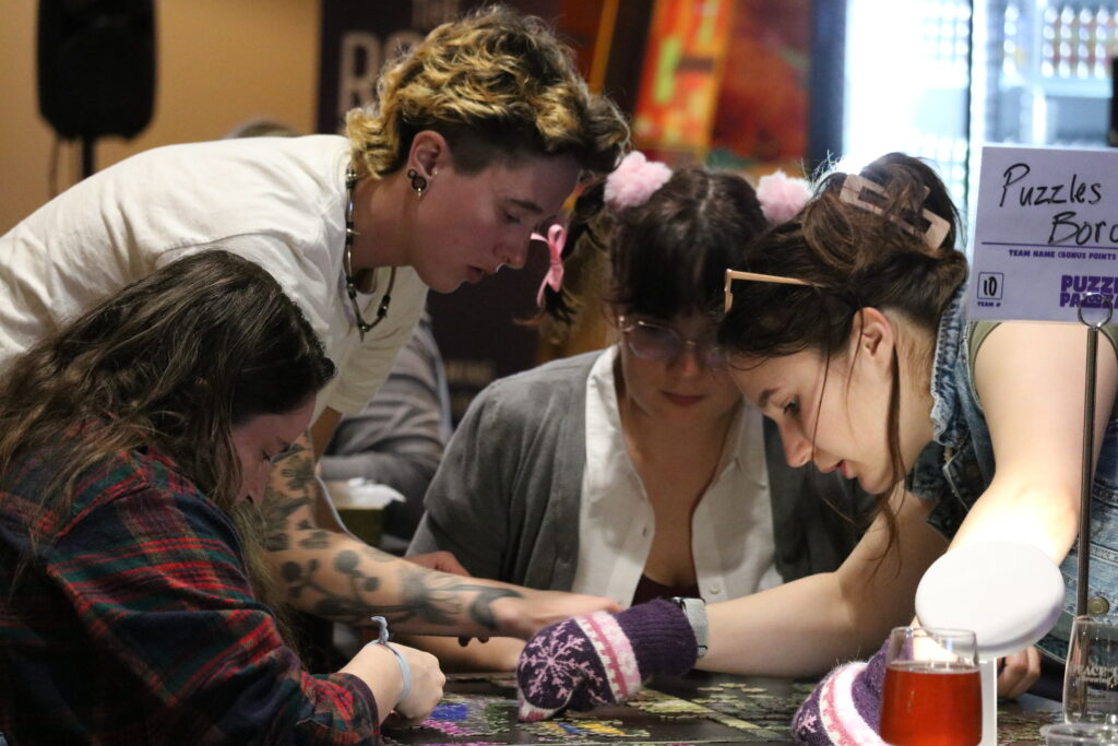 A group of people hunched over a table as they solve a jigsaw puzzle. One of them is has a mitten over one hand.