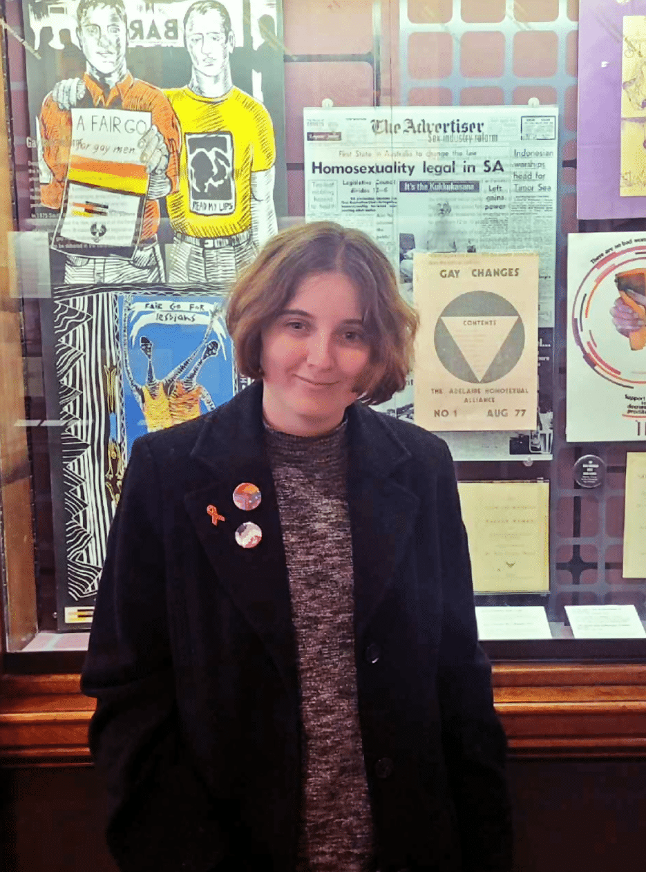 A person with a cropped bob wearing a dark overcoat affixed with several buttons stands in front of a glass case. In the case are newspaper clippings, artworks, flyers, and certificates.