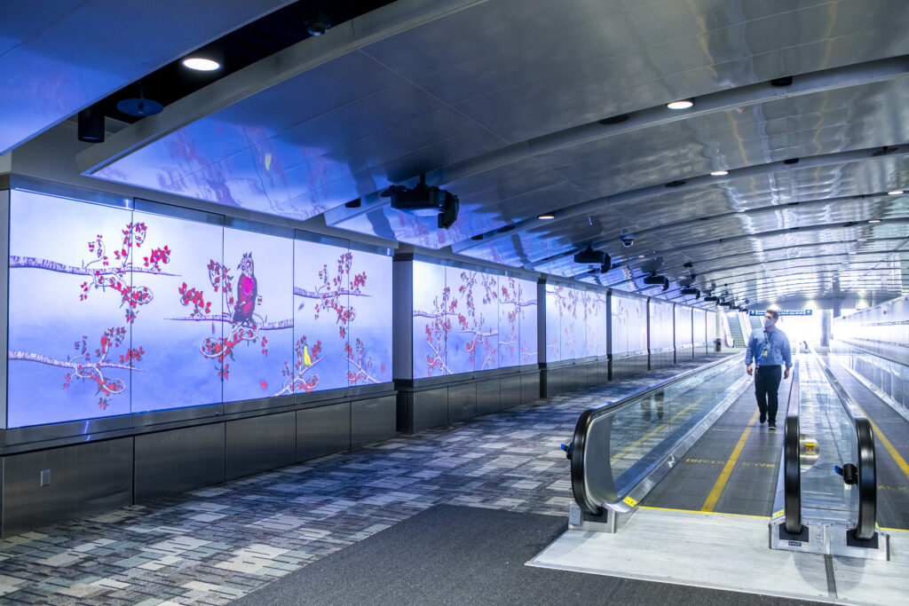 A long airport tunnel with screens on the side projecting artwork depicting tree branches with leaves and bird-like creatures. There is a person on the walkway looking at the art.