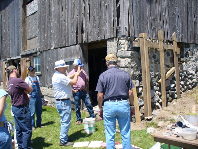 A group of people stand by an old wooden barn looking at its stone foundation. As others listen, one of them is gesturing with their hands.