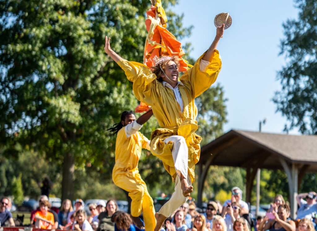 Two people in yellow outfits performing outside in front of children and adults. They leap joyously through the air with smiles on their faces.