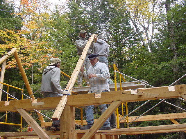 Four people stand on wooden beams as they build and assemble wooden rafters of a barn.