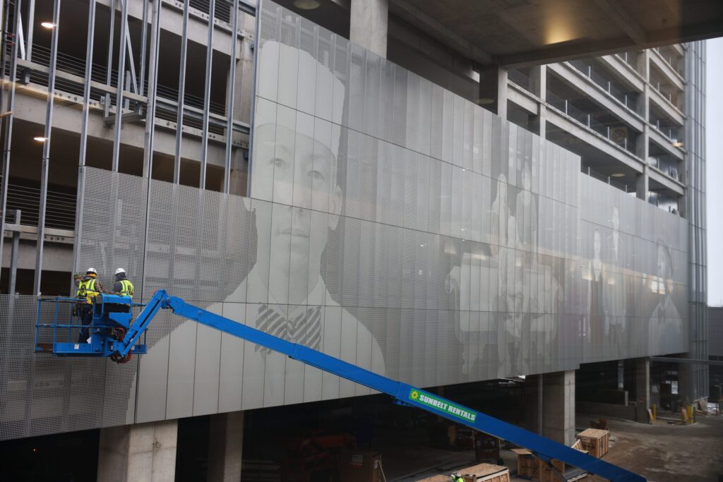 Two people on a tall construction lift as they install metal grids on a parking ramp. The metal grid depict photographs of people as part of an art installation.