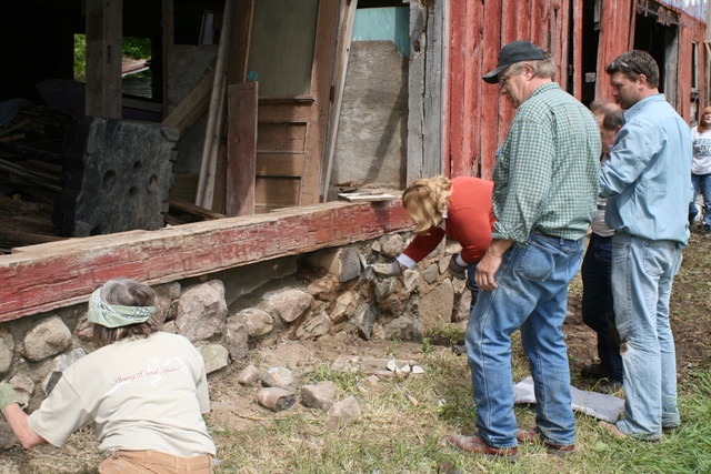 A group of people examining the stone foundation of an old wooden building. Some of them are standing, while a couple of them are looking at the stones more closely by leaning in or sitting on the ground.