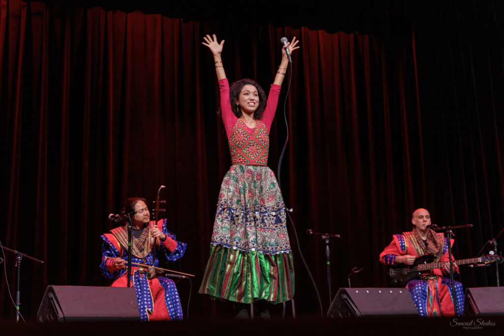 A woman wearing a colorful dress holds her hands above her head as two musicians playing instruments sit behind her on a stage.