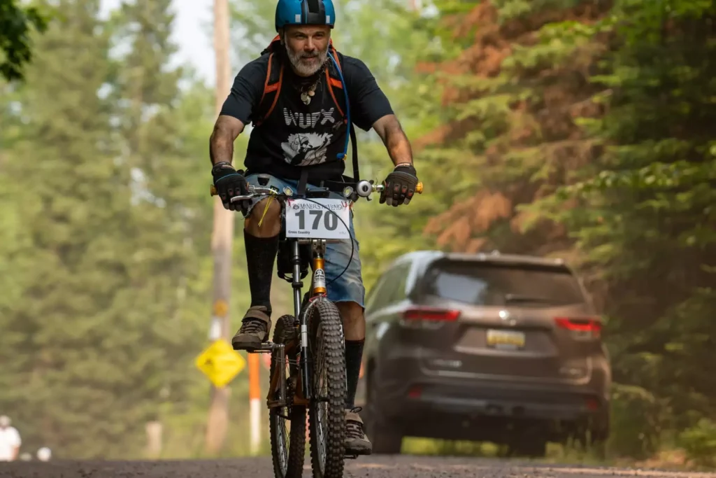 A person with a bright blue helmet and white beard looks determined as they pedal a bike down a gravel road. They are wearing a backpack, cutoff jeans, black gloves and a black t-shirt.