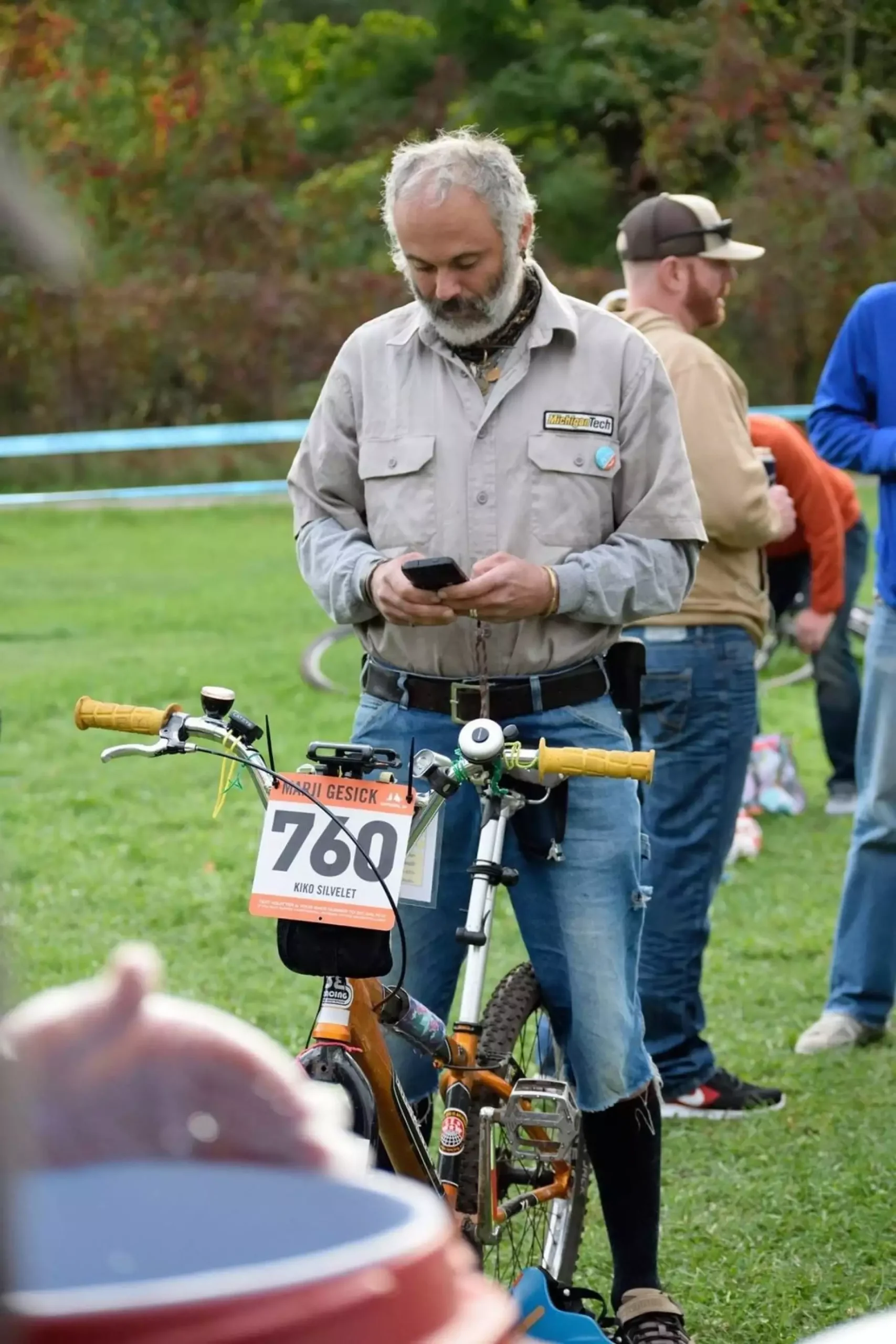 A person wearing jean cut-offs and a grey collared button up straddles a bike in a grassy field looking intently down at a phone in their hands.