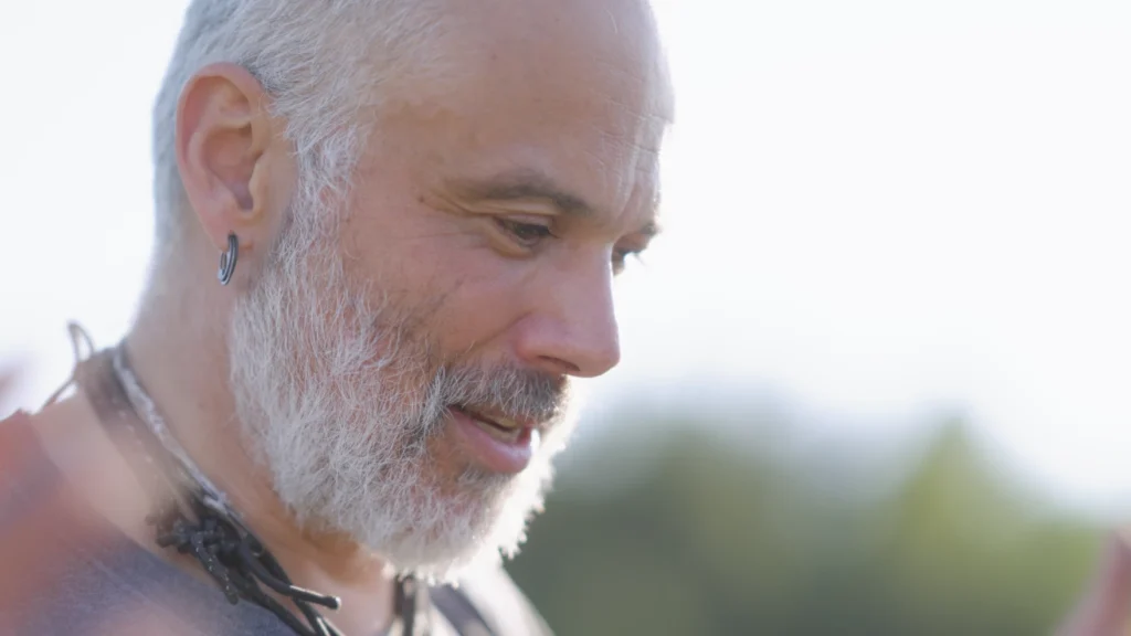 A close-up image of a person's face in profile. They have white hair and a white beard and a small hoop earring. They are softly smiling at something below.