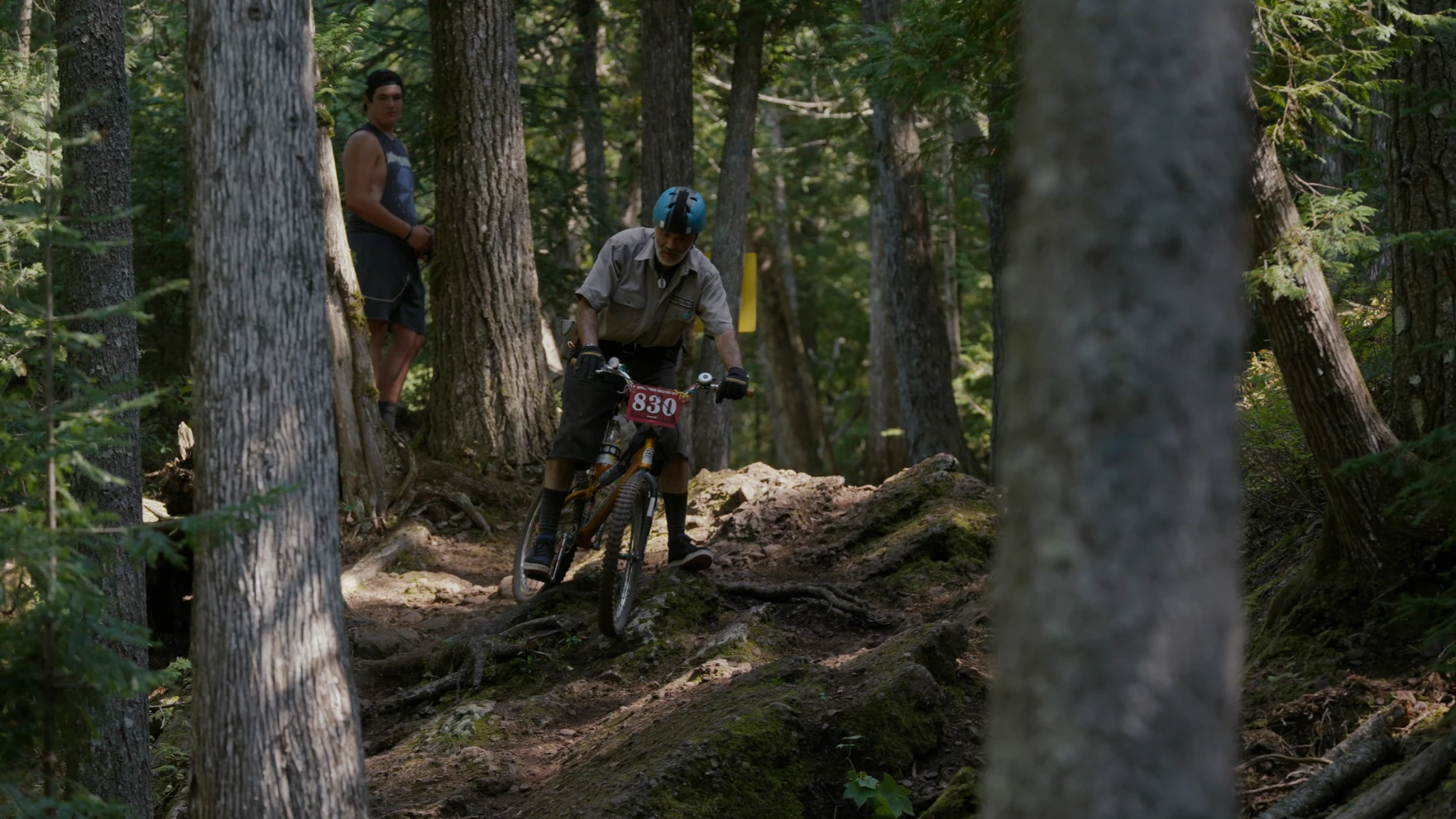 Shot through tall trees, a person attempts to navigate over thick roots on a bike. They are wearing a bright blue helmet and the bike has a tag with the number "830" on the front of its handle bars. Another person in a sleeveless shirt stands watching from behind.