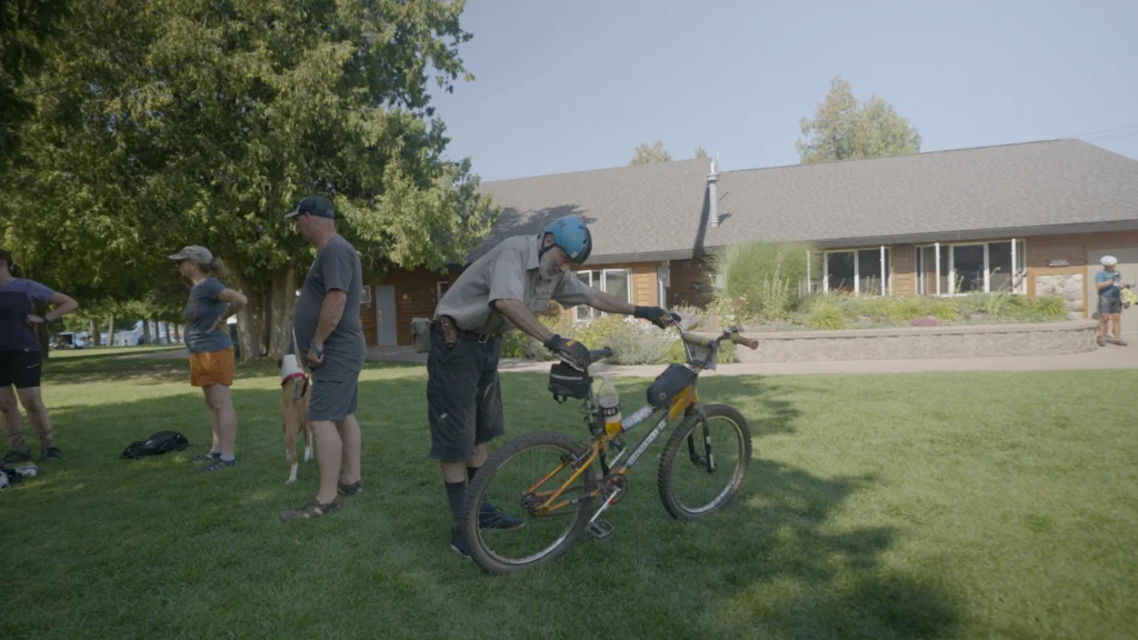 In a grassy meadow in front of a long one story long house a person leans over a bright yellow bike in the shadow of some trees - one hand on the seat, one hand on the handle bars. A couple of people and a dog on a leash stand facing the other direction to the left.