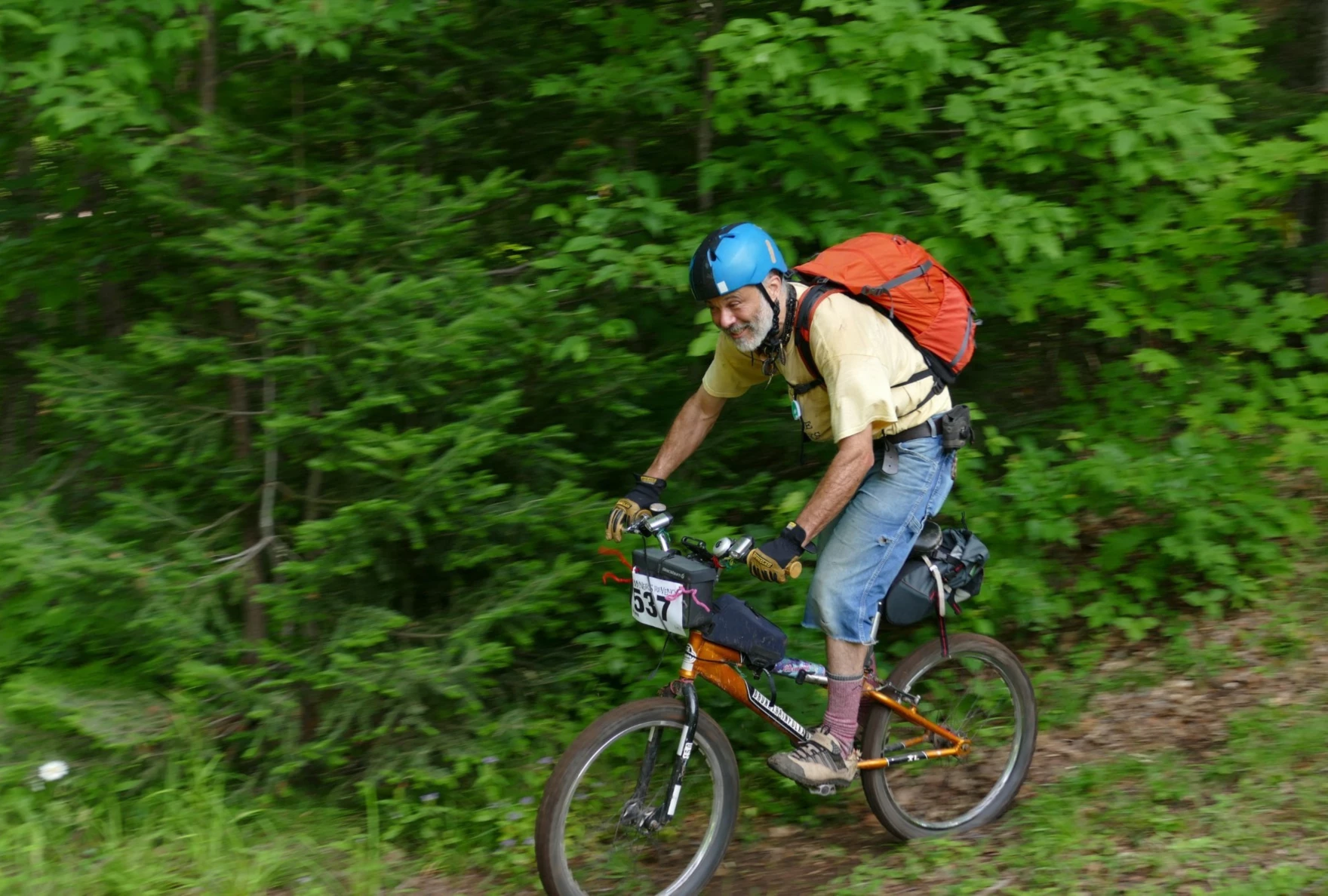 A person with a white beard wearing a bright orange backpack and bright blue helmet rides a bike standing up along a narrow dirt trail against a backdrop of deep green trees.