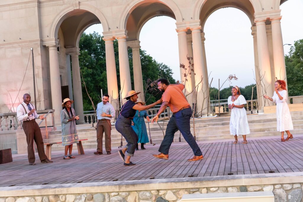 Six people standing on an outdoor stage with two people dancing in the center.