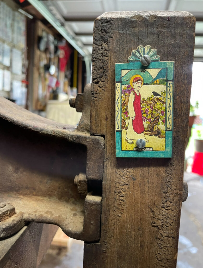 A piece of wood with an small painting nailed to it. The painting depicts a person wearing a red and white robe carrying a basket on their back and walking beside a vineyard.