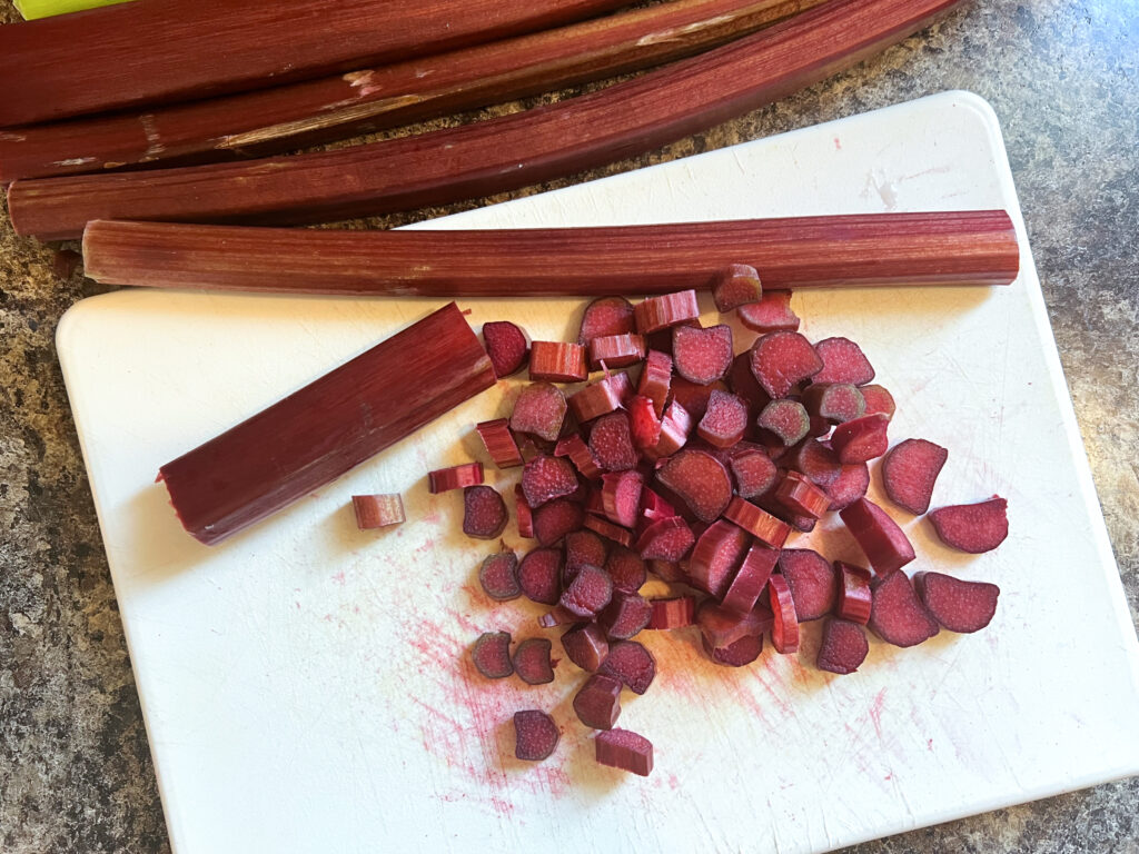 Chopped up pieces of deep red rhubarb on a white cutting board. There are long stalks of rhubarb beside the board.