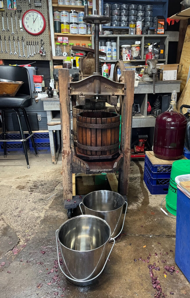 A wooden winemaking equipment with a metal press and wooden buckets. There are two metal buckets on the floor of what looks like a garage.