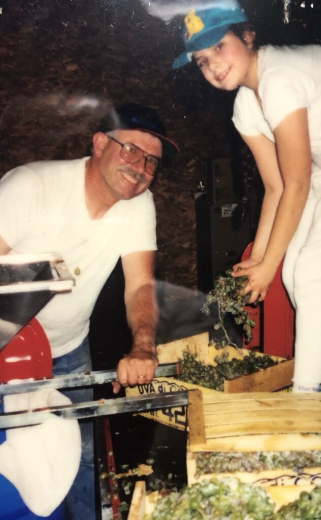 Two people in the process of making wine. One of them has a mustache and wearing glasses and a dark baseball hat. The other person is a young adult holding a handful of grapes in their hands.