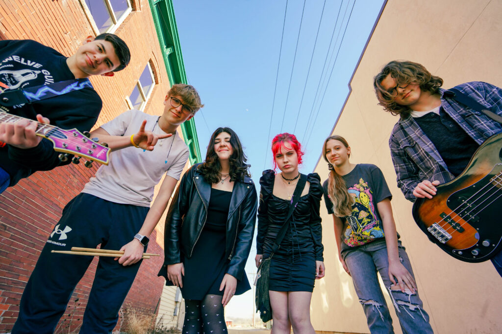 Six youth holding guitars and drumsticks posing outdoors.