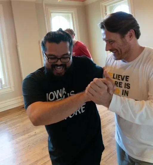 Two people dance with their hands clasped and arms around one another. They are smiling. One person in a black t-shirt, thick glasses and with a beard smiles with their mouth open looking down while their partner in a long sleeve white t-shirt looks at their partner.