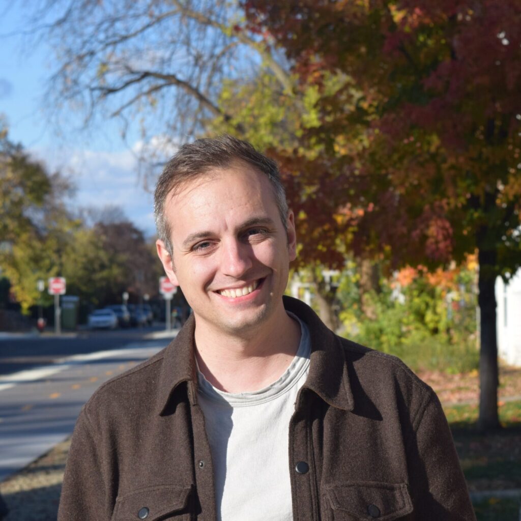 A person of light skin tone smiling. They have short dark grey hair and are wearing a white tshirt with a brown wool overshirt.