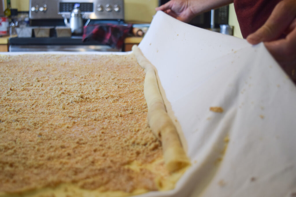 A large flat dough with a coarse powdery layer on top being rolled into a thin pipe-like shape.