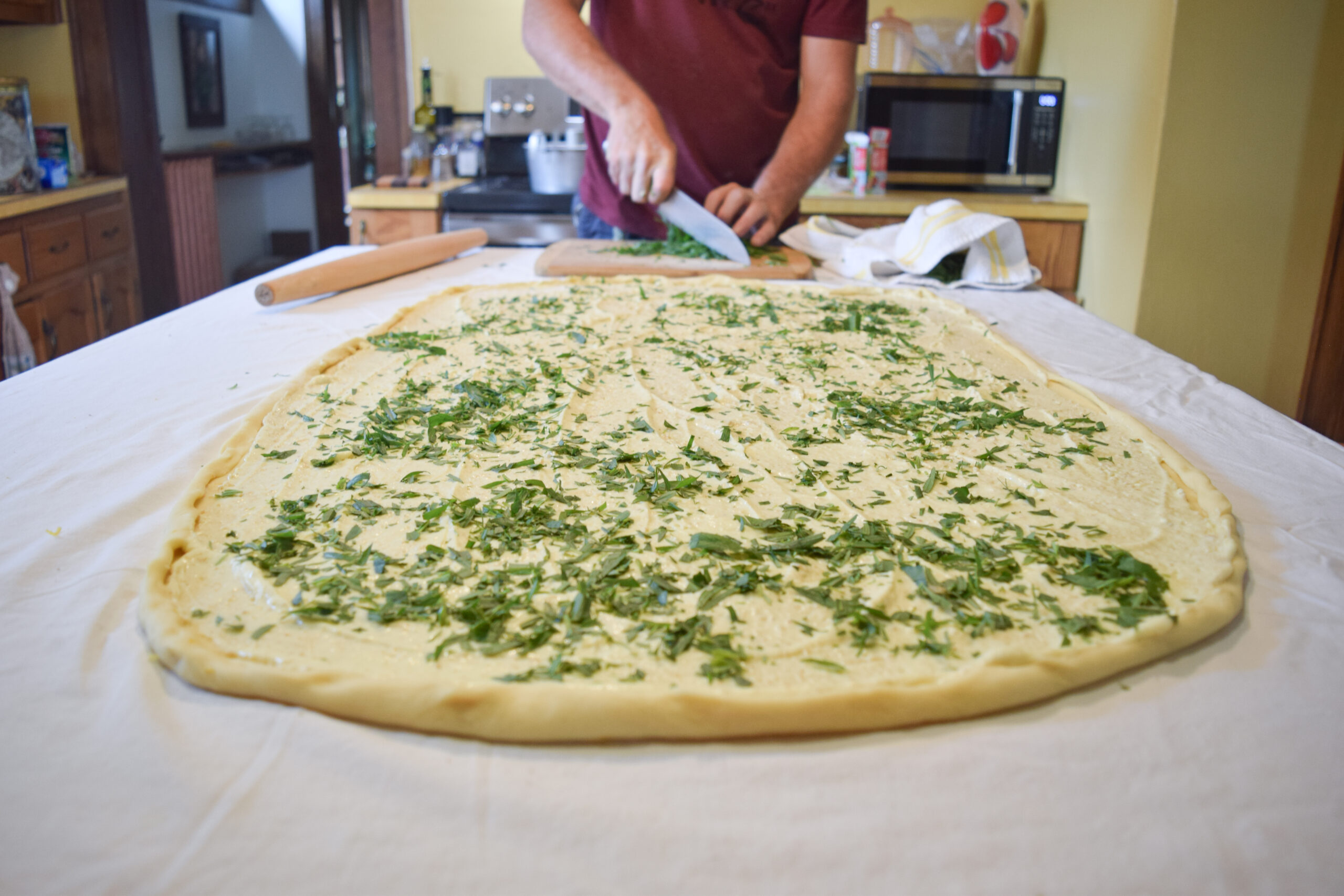 A large flat stretched dough with chopped green herb on top. At a distance, there is a person using a knife to chop green leaves on a wooden cutting board.