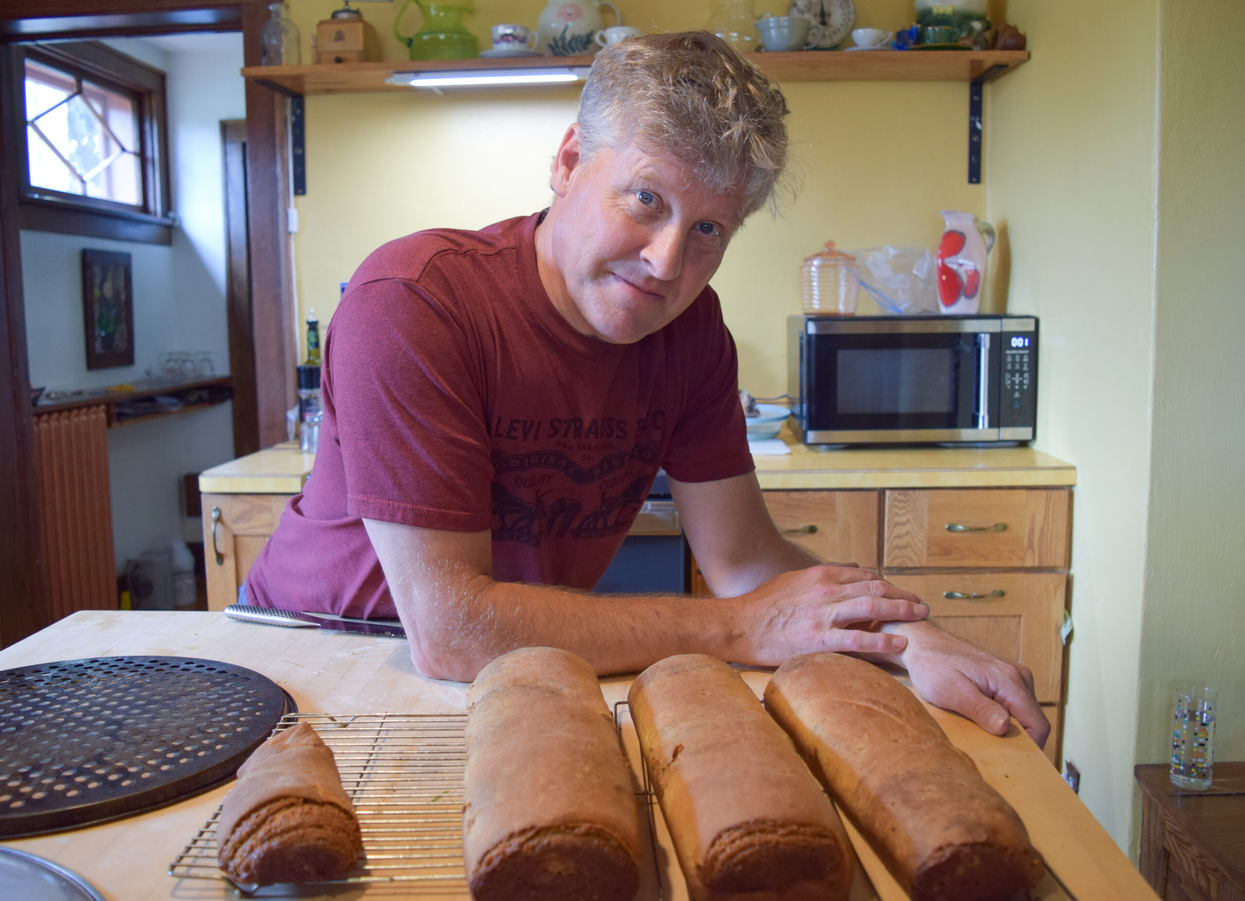 A person resting their arms on a wooden table and looking straight on. They has grey hair and are wearing a maroon tshirt. There are baked good in front of them on the table.