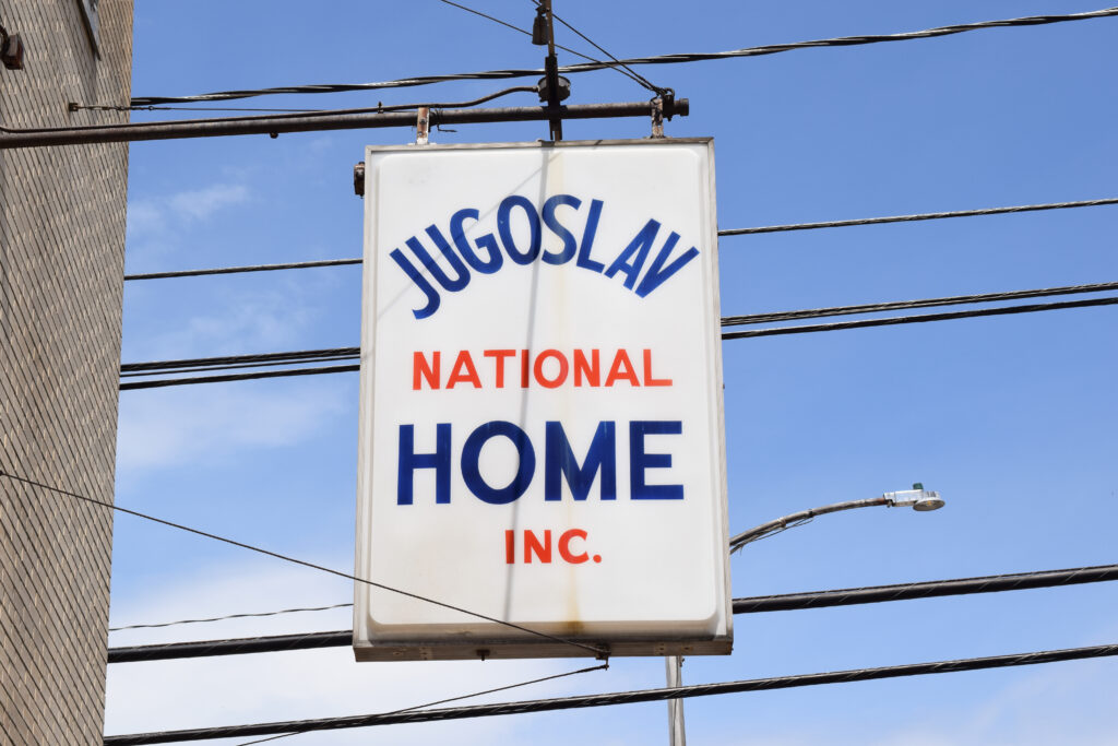 A white sign on a building with lettering in blue and red. It reads "Jugoslav National Home Inc."