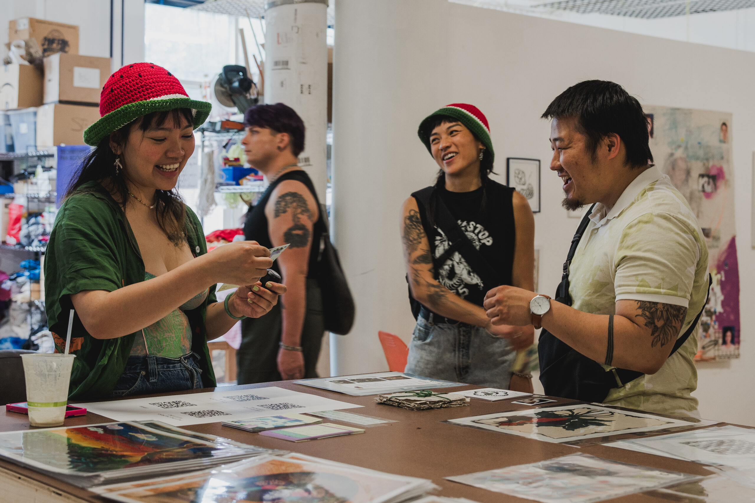 Three people stand around a table with various artworks on paper. They are all smiling or laughing. Two of them are wearing crocheted bucket hats made to look like watermelon.