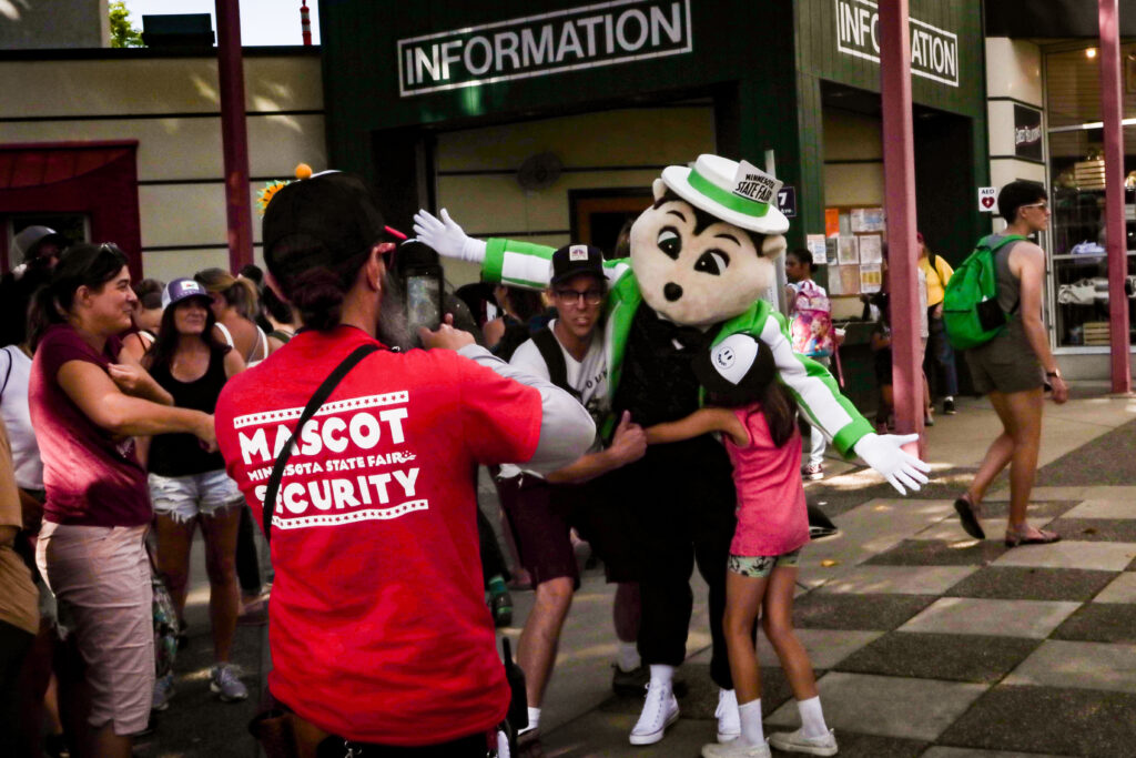 A person dressed in a mascot costume as a gopher wearing a green and white striped suit and boater hat, posing with fans as a member of the mascot security team takes a photo.