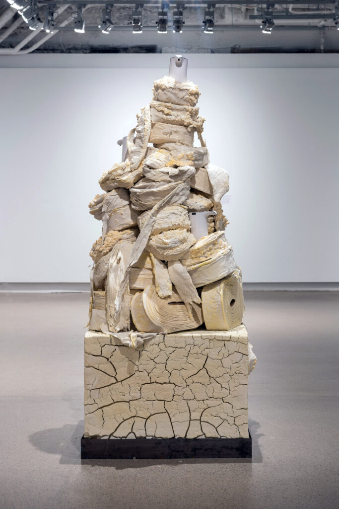 A sculpture made up of a stack of yellowing rolls of toilet paper and touchless soap dispensers standing in a gallery space.