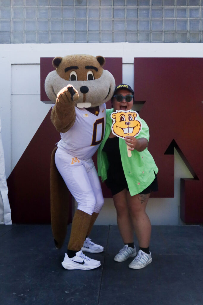 A person wearing black shorts and a green shirt holding a hand fan in the shape of a gopher's head, posing with a person in a mascot costume of a gopher wearing a football uniform.