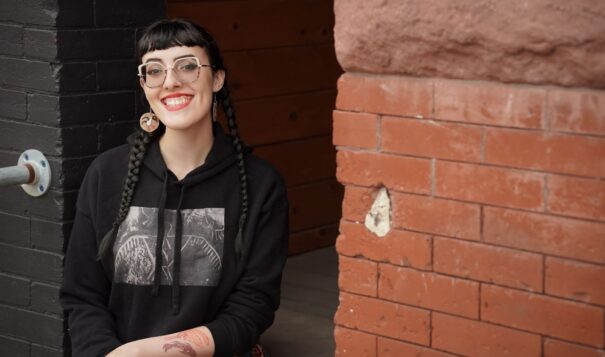 A person with two long black braids and bangs sits in a doorway next to a red brick wall smiling. They are wearing a black hoodie, large circular earrings, and large glasses.