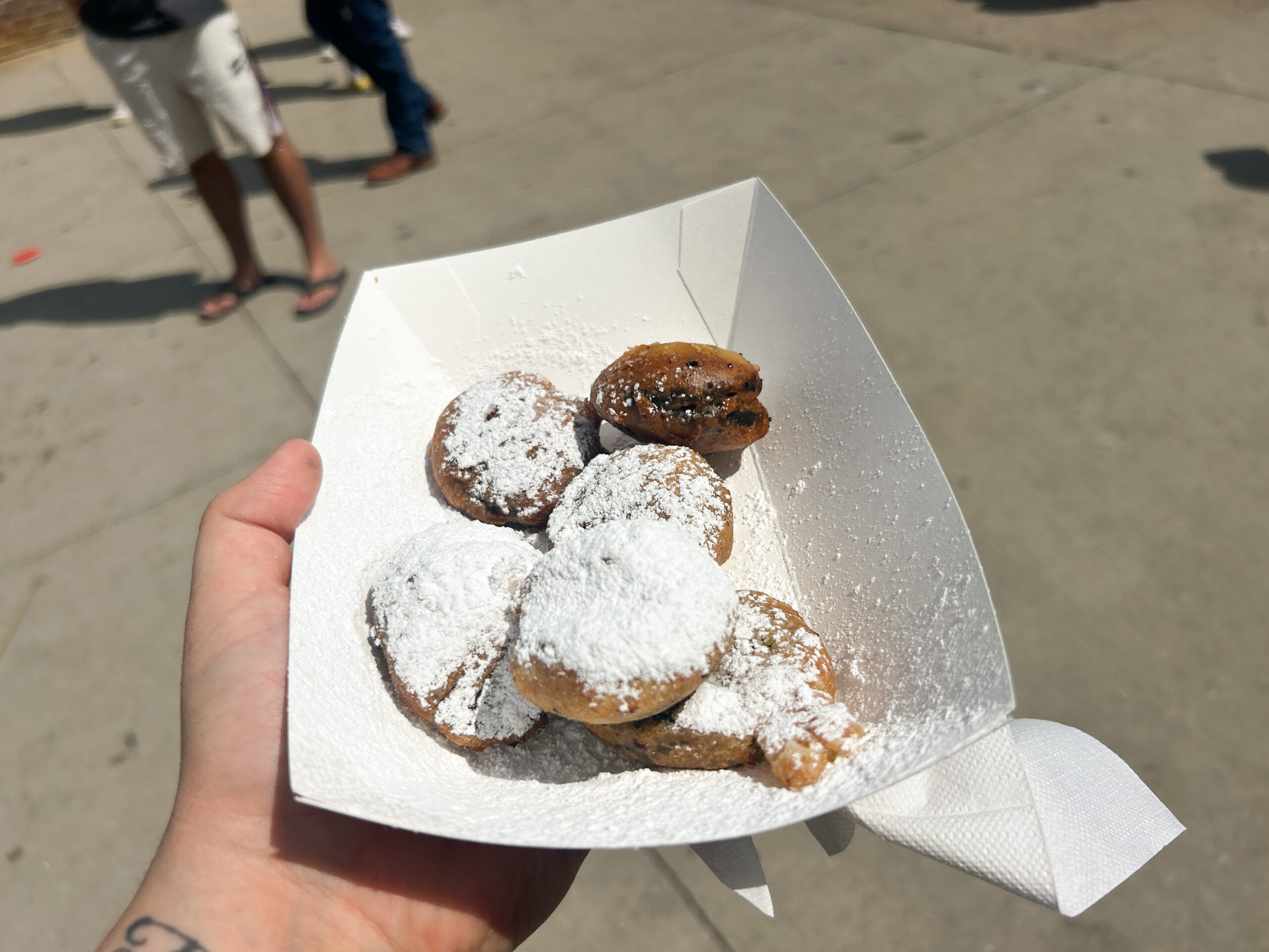 A hand holding a paper bowl with round, deep fried food item. There is powdered sugar sprinkled on top of them.