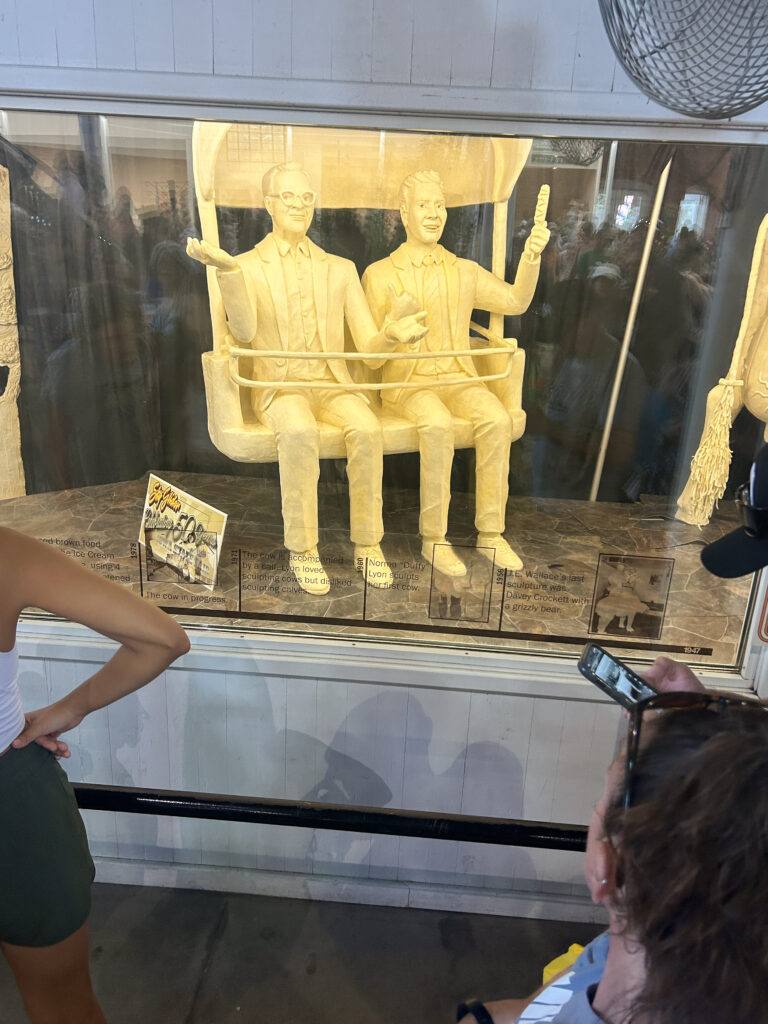 A sculpture made of pale yellow-colored butter depicting two full-size people sitting in a sky lift. They are in a display case with a window. There are people looking at the display.