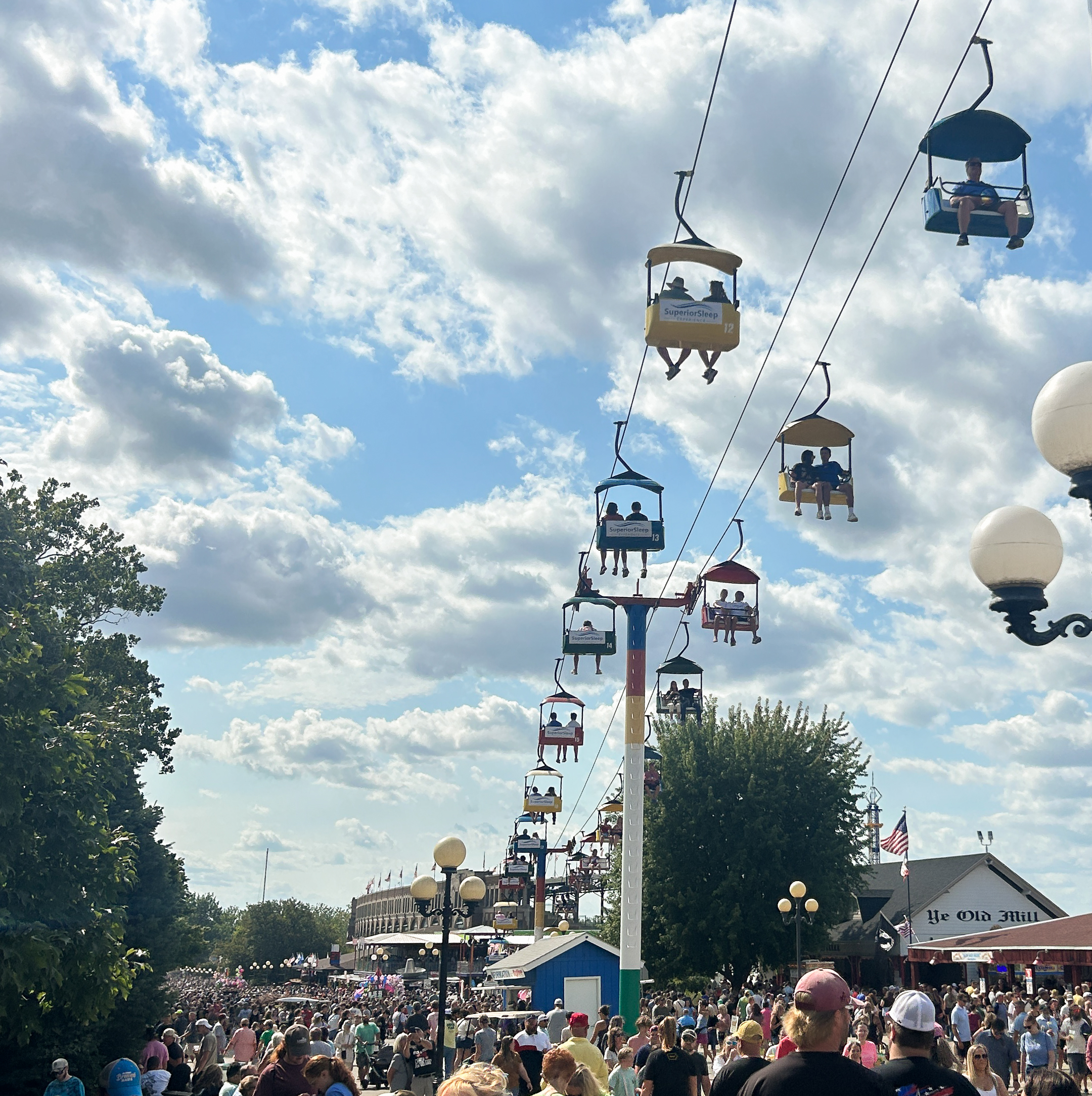 Crowds of people walking in an open area, as others sit in sky lifts above the crowd.