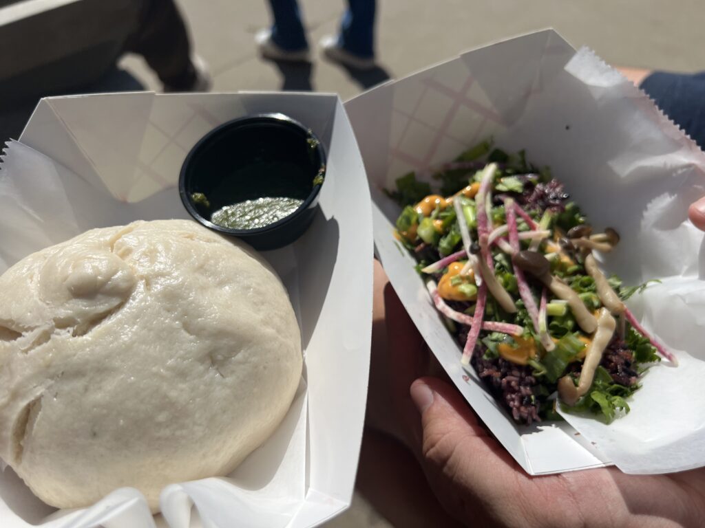 A large steamed bun with a green sauce, and a sticky rice dish with watermelon radish, cilantro, green onions, and mushrooms.