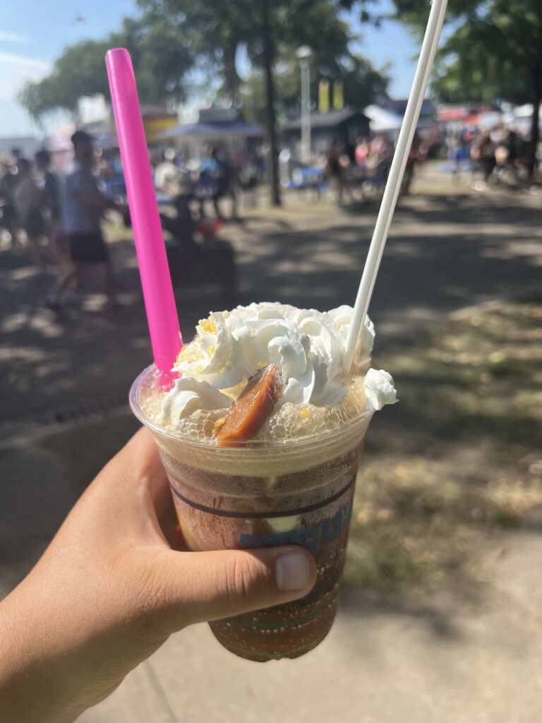 A plastic cup of cola with a scoop of yellow ice cream, whipped cream, and a small piece of caramel, with a pink straw and long white spoon sticking out of it.