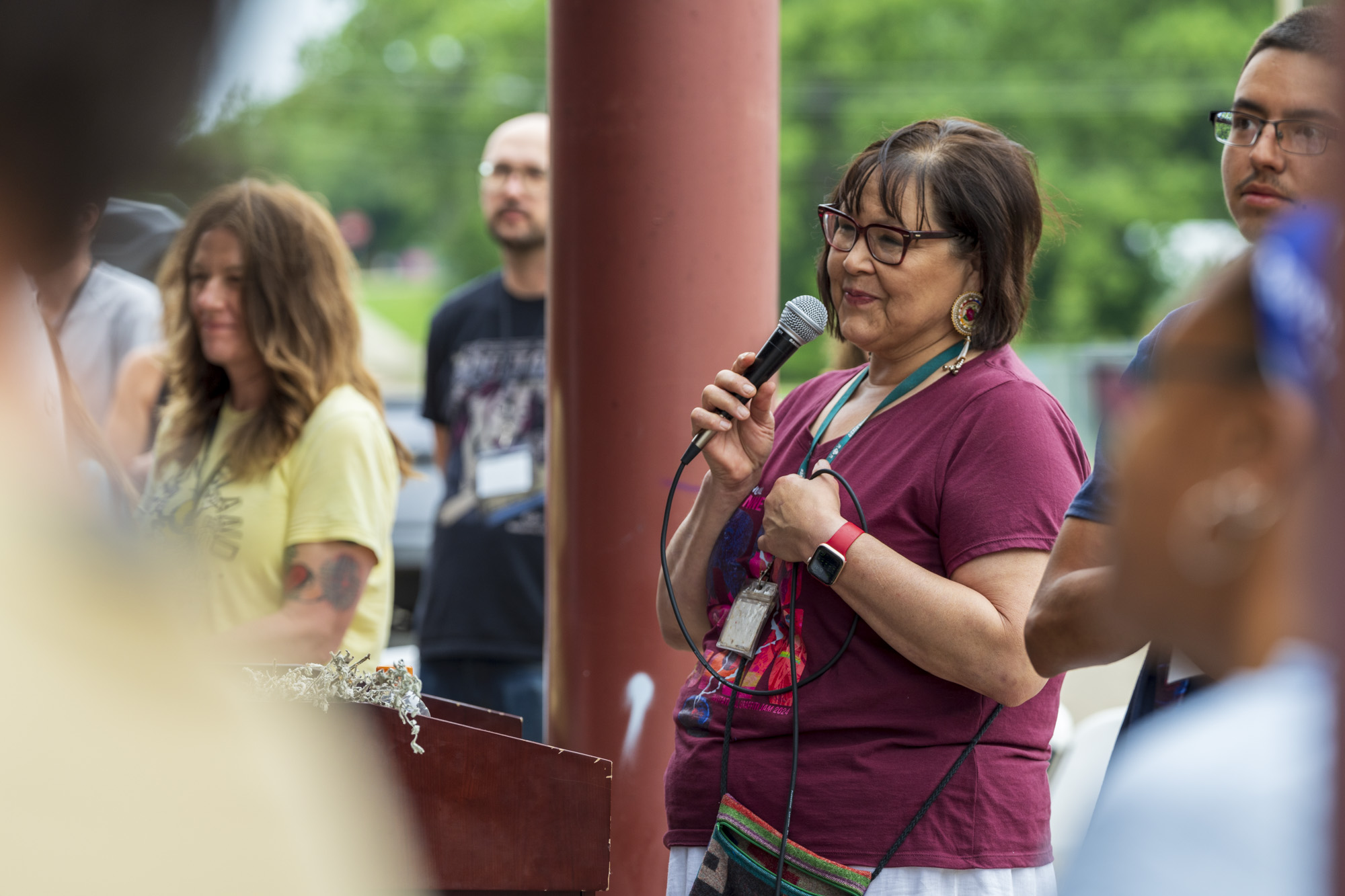 A person of medium light skin tone and dark shoulder-length hair holding a mircrophone. They are wearing a maroon tshirt and dark-rimmed glasses.