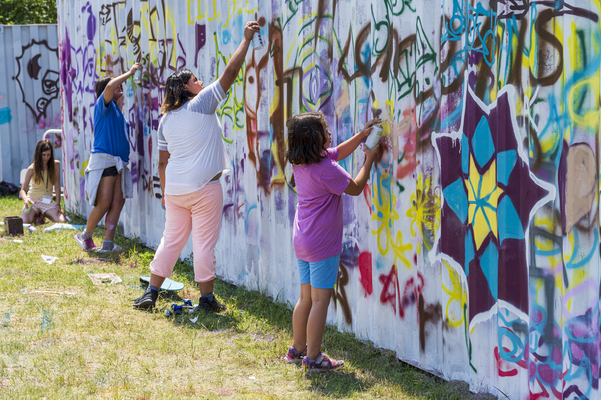 Kids of varying ages hold spray paint cans and add to graffiti art on a wall in front of them.