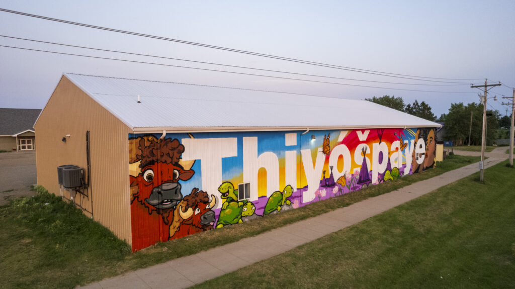 A mural on a side of a building with bold white lettering that spells "thiyóšpaye" and paintings depicting animals, nature and people.