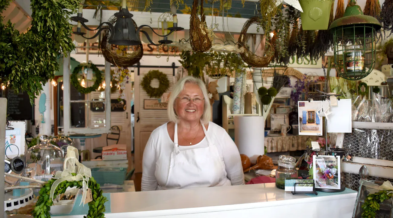 A person with shoulder-length white hair wearing a white shirt and white apron stands smiling behind a counter that comes to above their waste. In the background wreaths of many different natural elements and textures hang from the ceiling and walls including various light fixtures making for a cluttered, homey atmosphere.